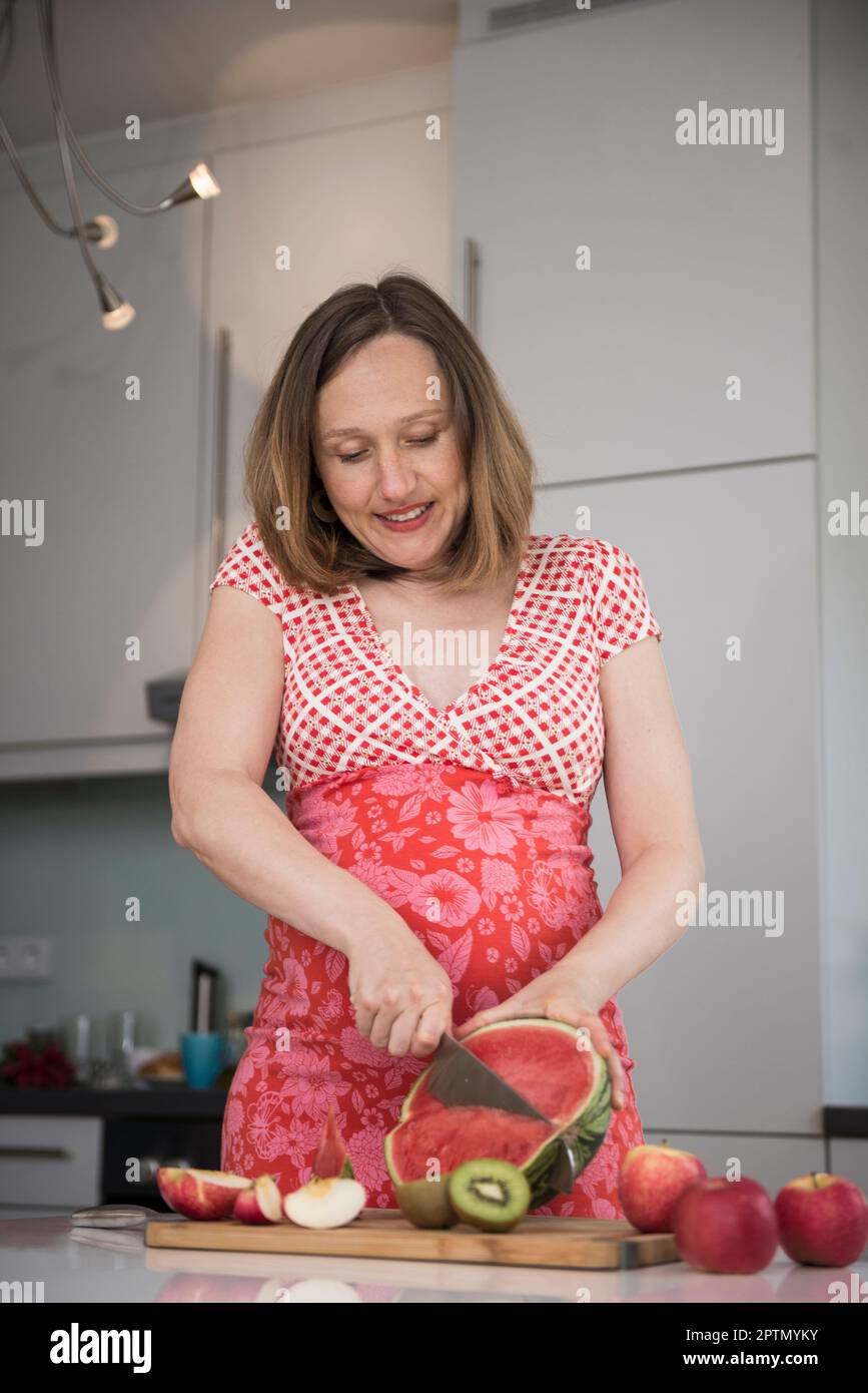 Femme enceinte coupant des tranches de pastèque dans la cuisine, Munich, Bavière, Allemagne Banque D'Images
