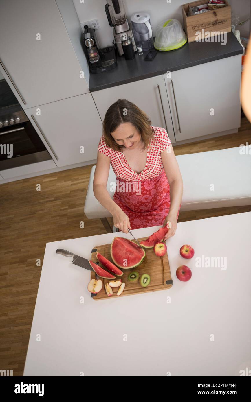 Femme enceinte coupant des tranches de pastèque dans la cuisine, Munich, Bavière, Allemagne Banque D'Images