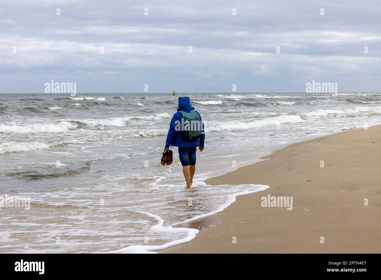 Un homme habillé d'une veste bleue marchant avec ses pieds nus sur le sable au bord de la mer Baltique. Plage vide hors saison, temps venteux, W Banque D'Images