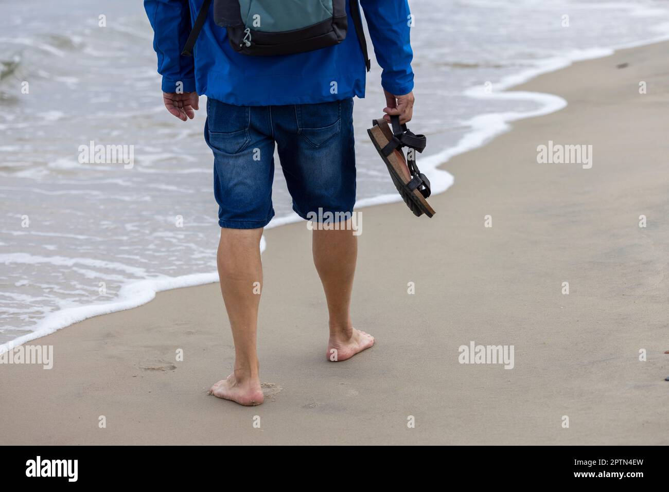 Un homme habillé d'une veste bleue marchant avec ses pieds nus sur le sable au bord de la mer Baltique. Plage vide hors saison, temps venteux, W Banque D'Images