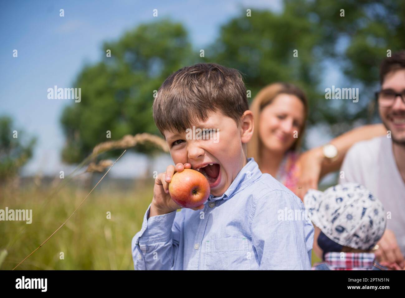 Petit garçon mangeant une pomme sur la prairie dans la campagne avec la famille en arrière-plan, Bavière, Allemagne Banque D'Images