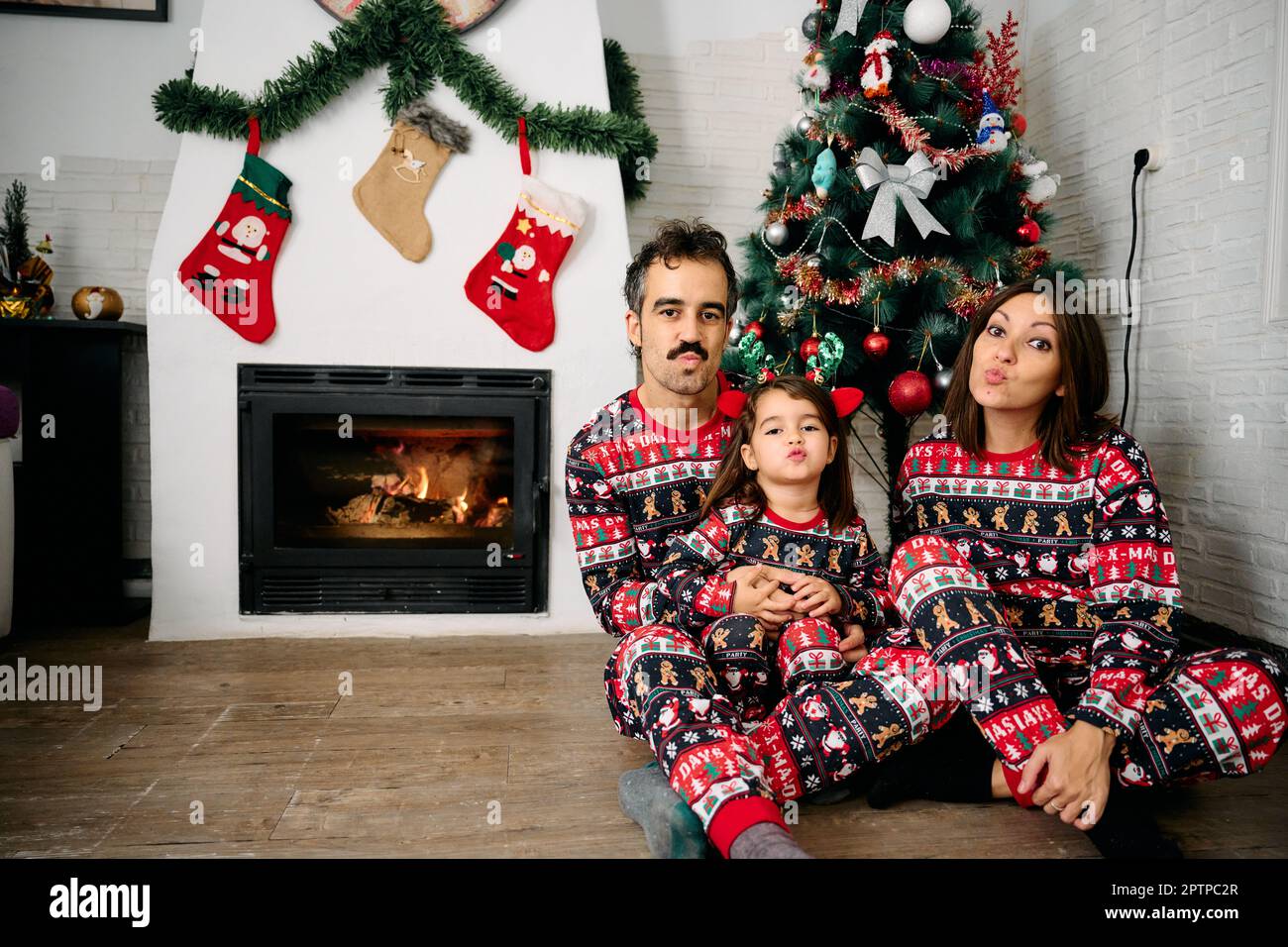 Une famille, avec son père, sa mère et sa fille, portant un pyjama de Noël assorti, prenant des photos de famille devant la cheminée et un tr de Noël Banque D'Images