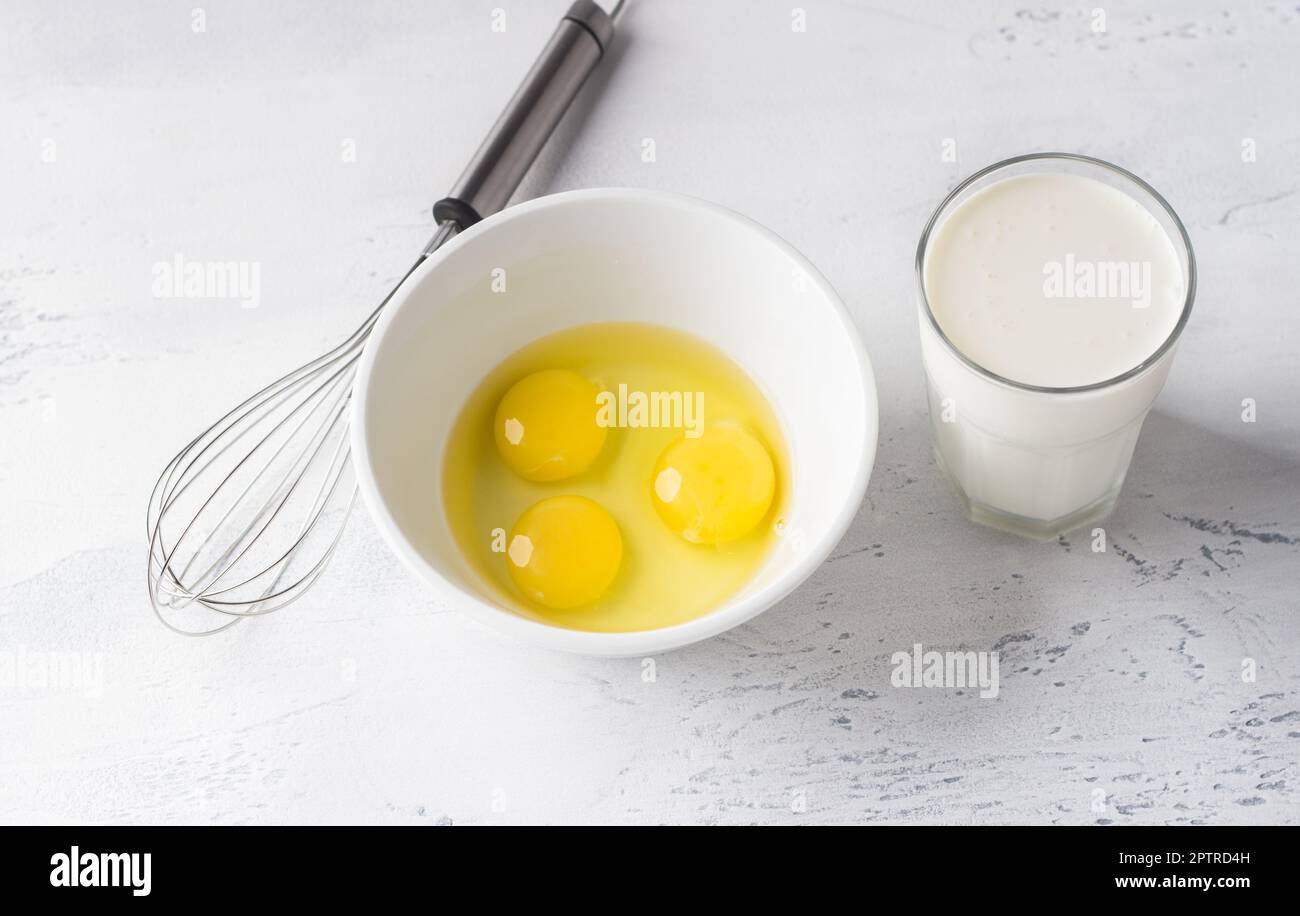 Bol blanc avec trois œufs cassés sans coquille, un verre de crème ou de lait et un fouet pour fouetter sur fond gris clair, vue du dessus. Cuisine homem Banque D'Images