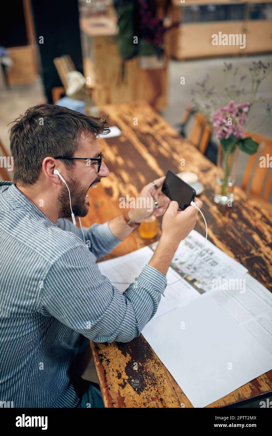 Jeune homme attrayant joyeux assis seul dans un café avec un casque à l'aide du téléphone, appels vidéo avec un ami. Café moderne. Banque D'Images