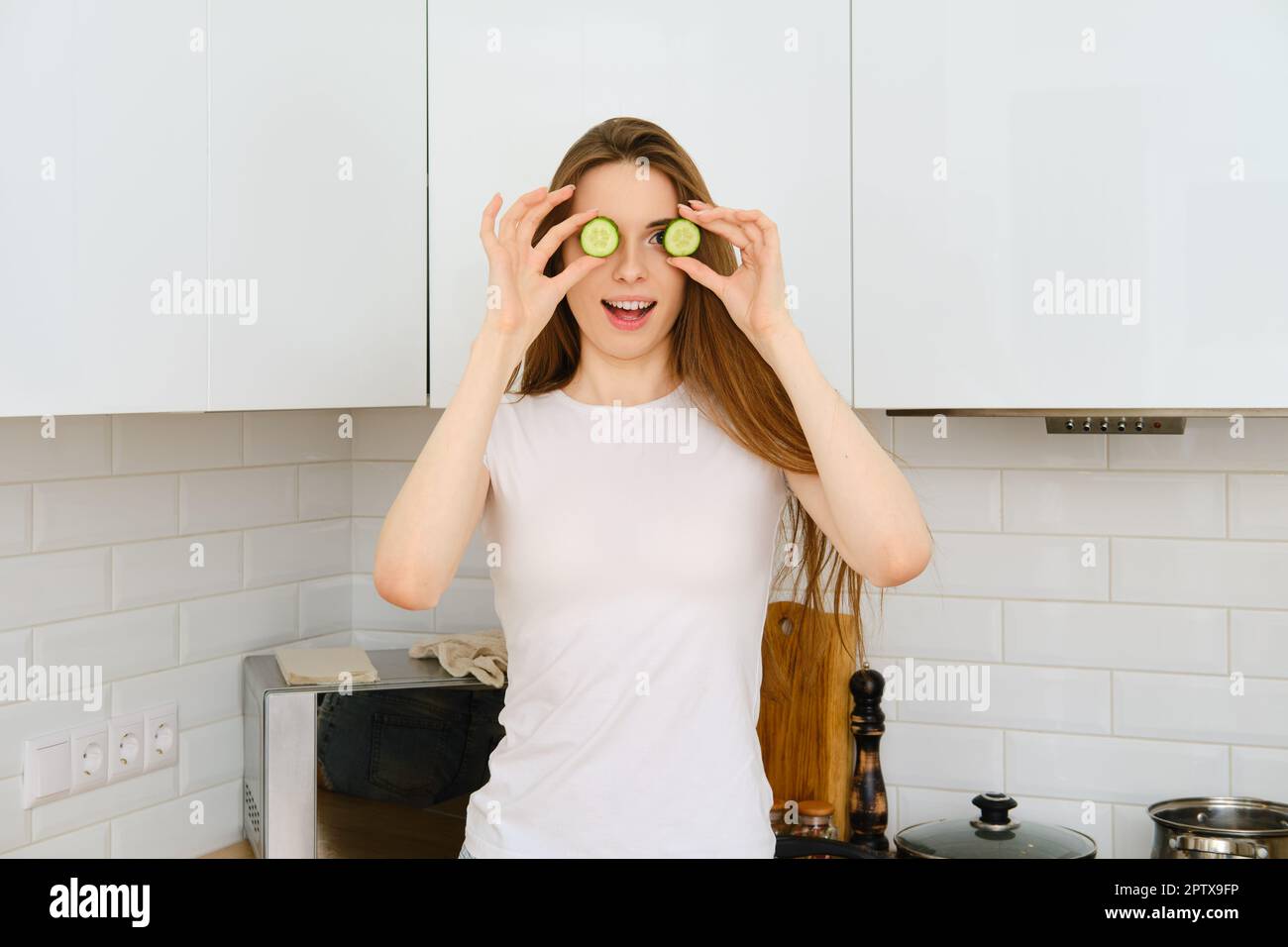 Drôle de photo de la jeune femme joyeuse en t-shirt blanc couvrant les yeux avec des tranches de concombre et souriant à l'appareil photo, la femme s'amusant tout en cuisinant dans moderne Banque D'Images