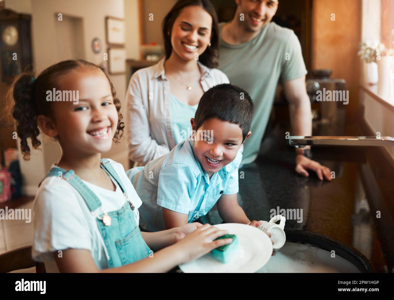 Jeunes parents heureux de race mixte laver les plats avec leur fils et leur fille dans la cuisine à la maison. Petit frère et soeur aidant leur mère et Banque D'Images