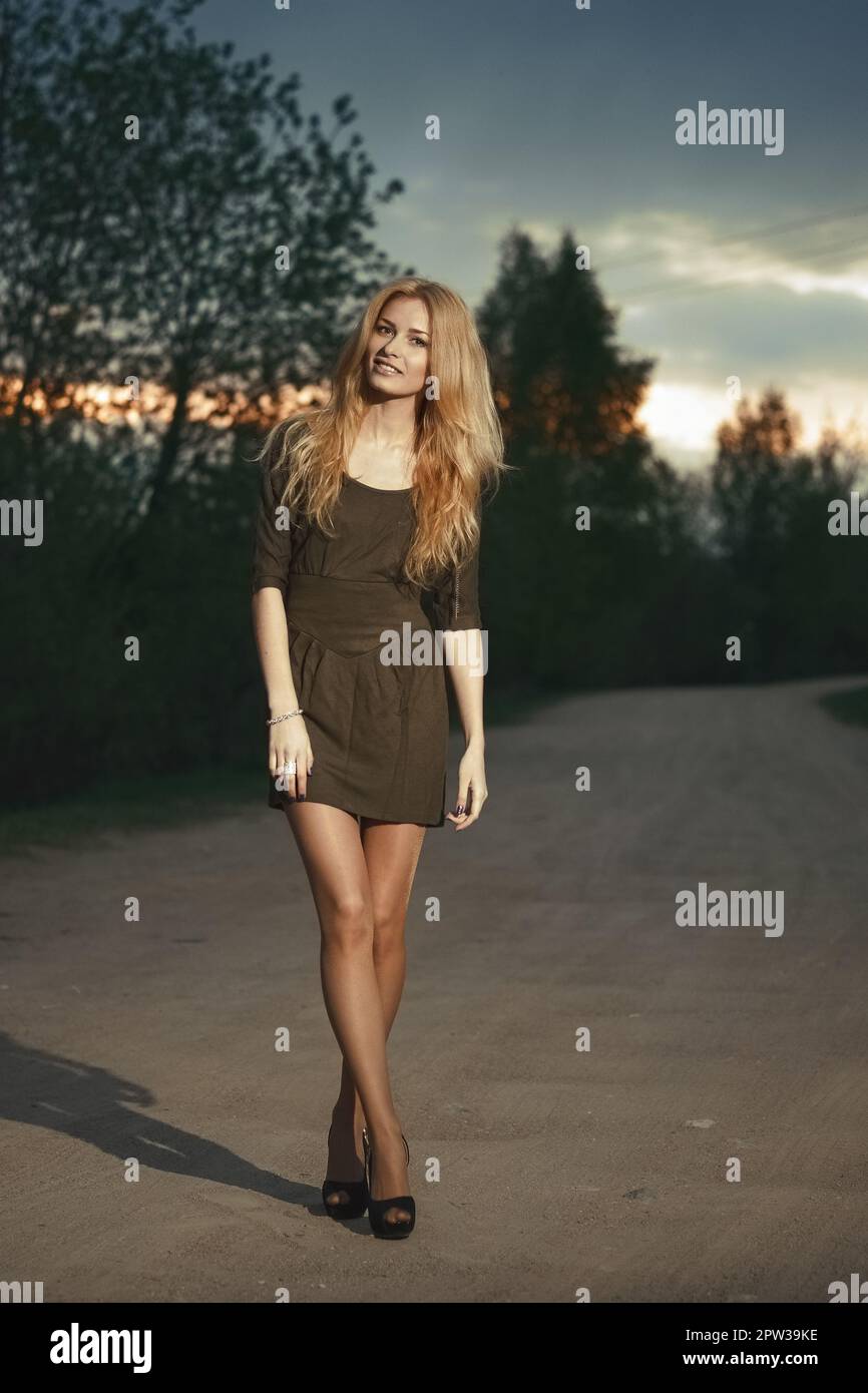 Portrait d'une jolie femme blonde à l'heure du coucher du soleil sur la route. De longues jambes et des talons hauts. Banque D'Images