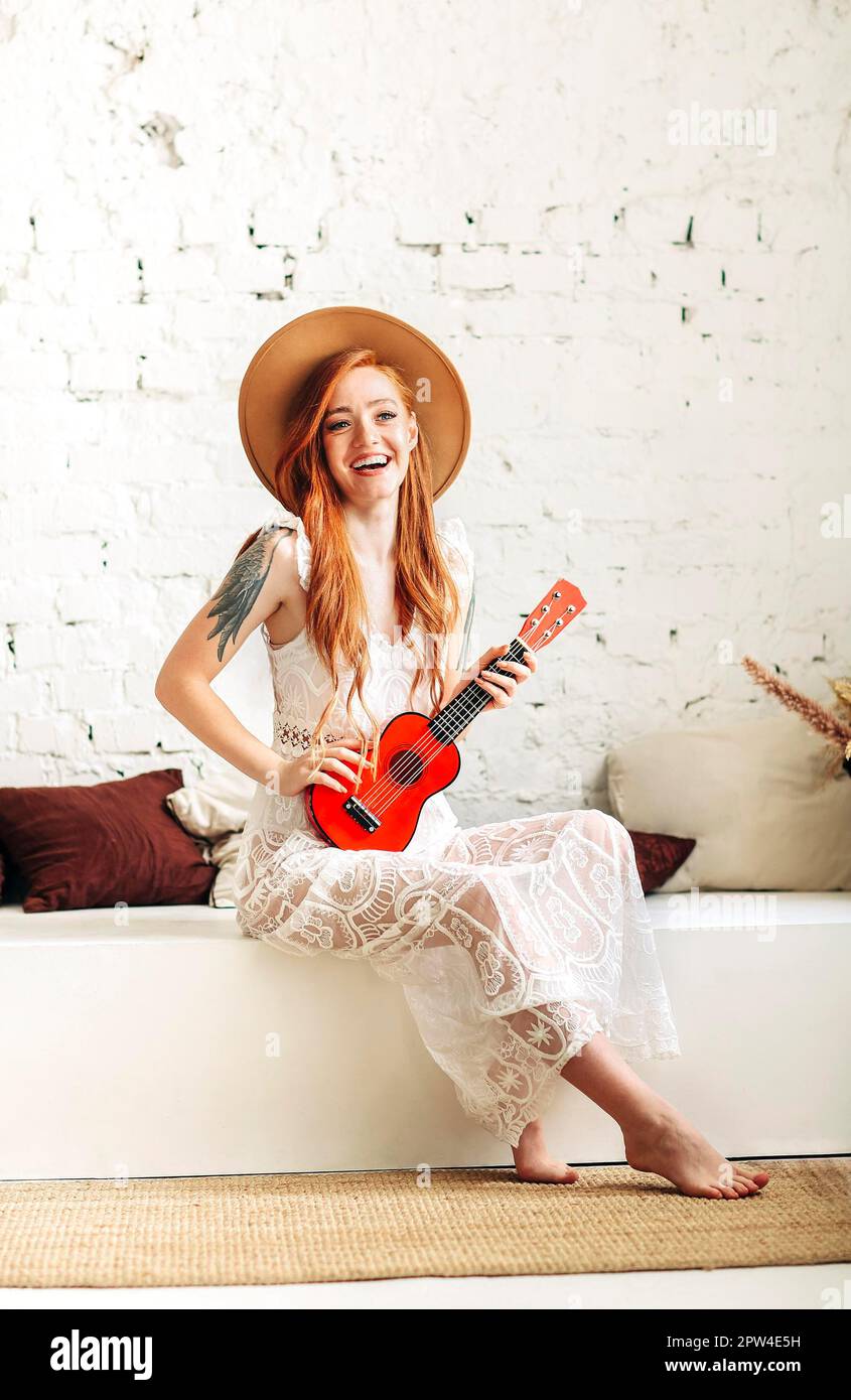 Plein corps bonne jeune femme dans la robe de boho et chapeau souriant et jouant ukulele tout en étant assis sur le banc contre le mur de brique blanche à la maison Banque D'Images