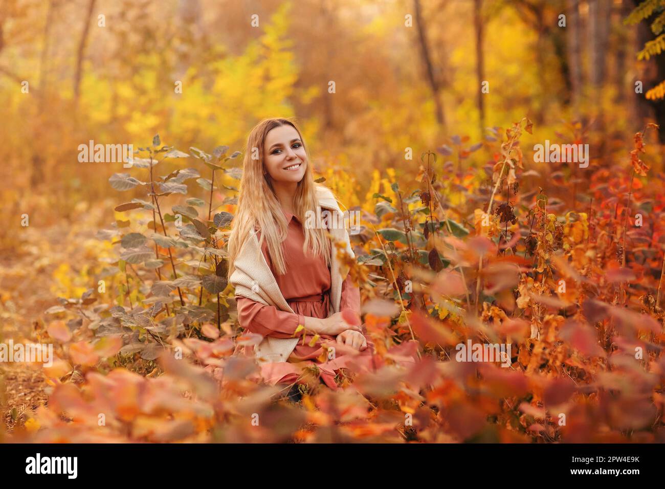 Belle jeune femme blonde portant une robe longue de style boho tout en étant assise avec un look pensif dans l'herbe haute en forêt d'automne décorée dans le rouge et le jaune Banque D'Images