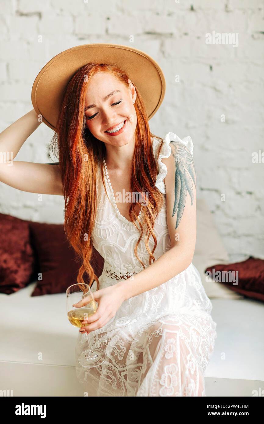 Bonne jeune femme en robe boho et chapeau en dégustant du vin avec le sourire tout en étant assise sur le banc contre le mur de briques blanches Banque D'Images