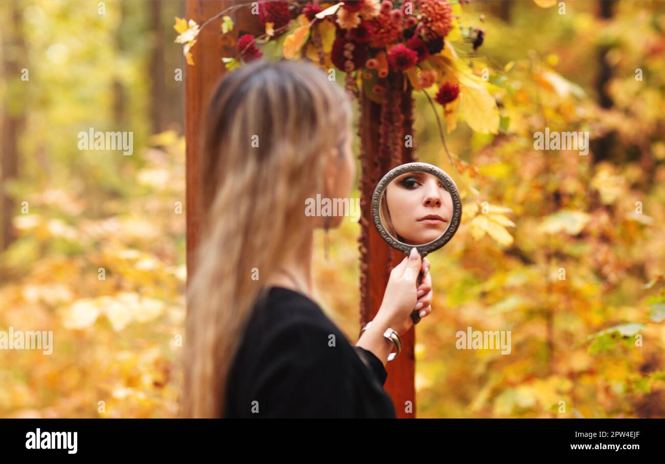 Photo pittoresque de la jeune fille belle avec maquillage du soir portant le costume de sorcière noir tenant le miroir et le regardant pendant la séance photo d'halloween Banque D'Images