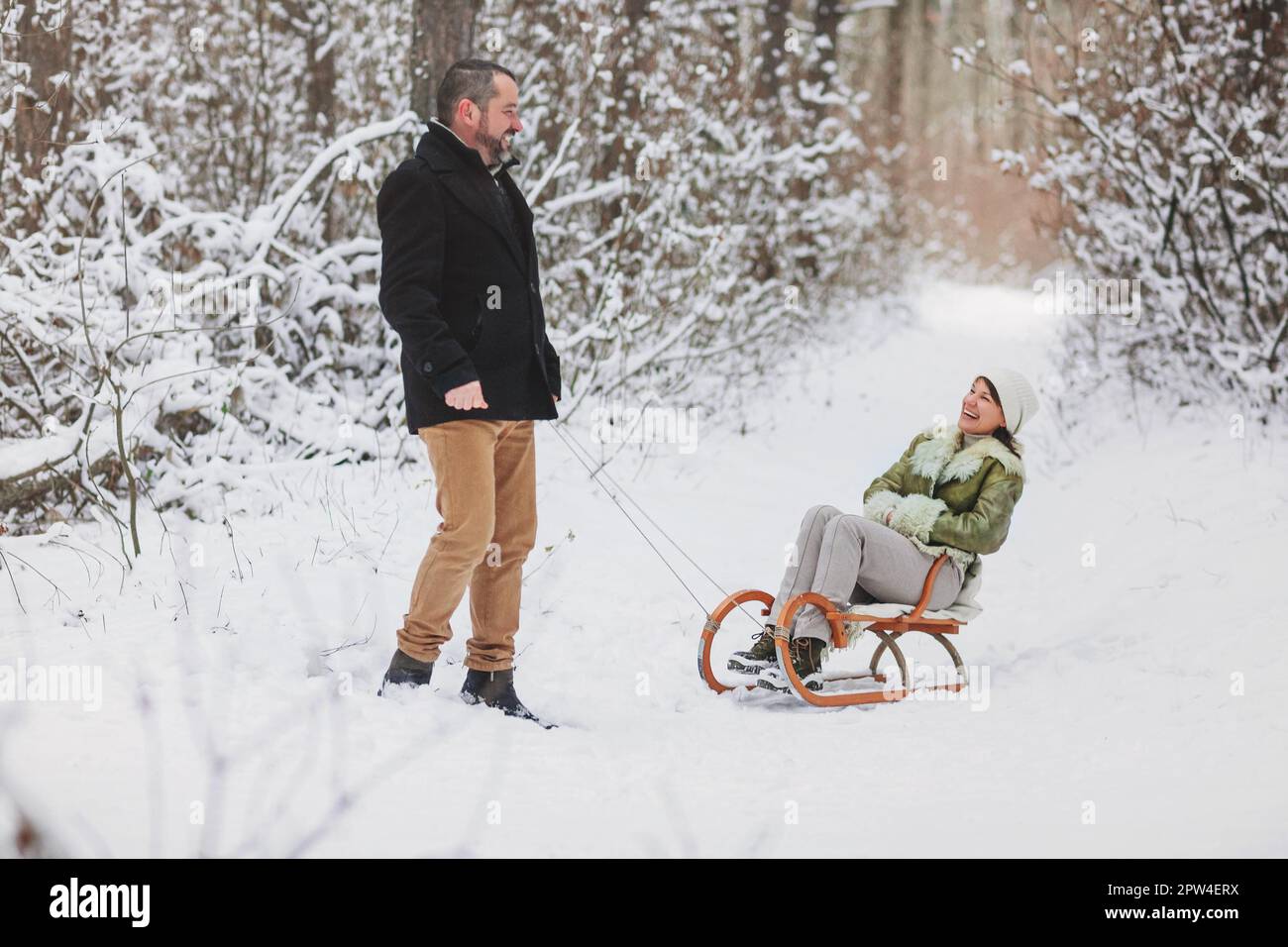Joyeux couple de famille mûr et amusant en train de traîner dans le parc d'hiver, en riant et en s'amusant ensemble, femme d'âge moyen positive assise sur un traîneau pendant qu'elle Banque D'Images