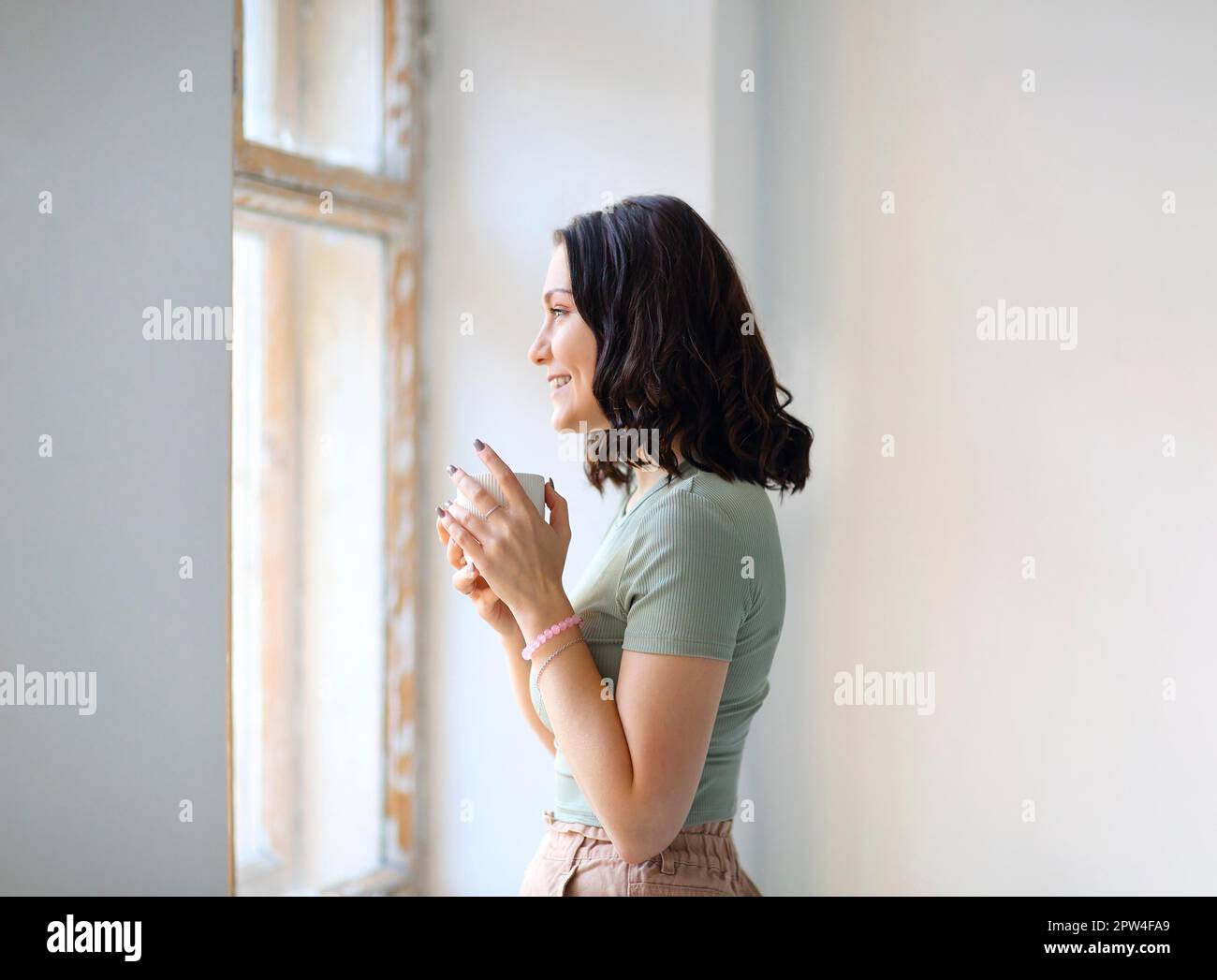 Photo latérale d'une jeune brune gaie debout à l'intérieur avec une tasse de thé tout en regardant la fenêtre avec un doux sourire en regardant le temps dehors. Banque D'Images