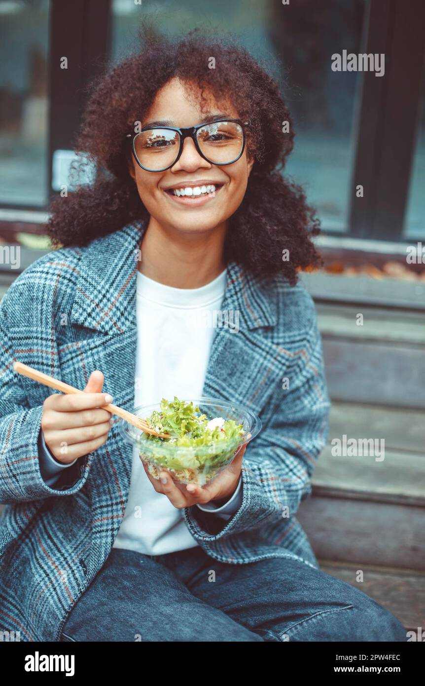 Femme afro-américaine heureuse travaillant au bureau en train de manger de la salade et de sourire à la caméra tout en étant assise sur le banc dans le parc à l'extérieur, foyer sélectif. Positif Banque D'Images