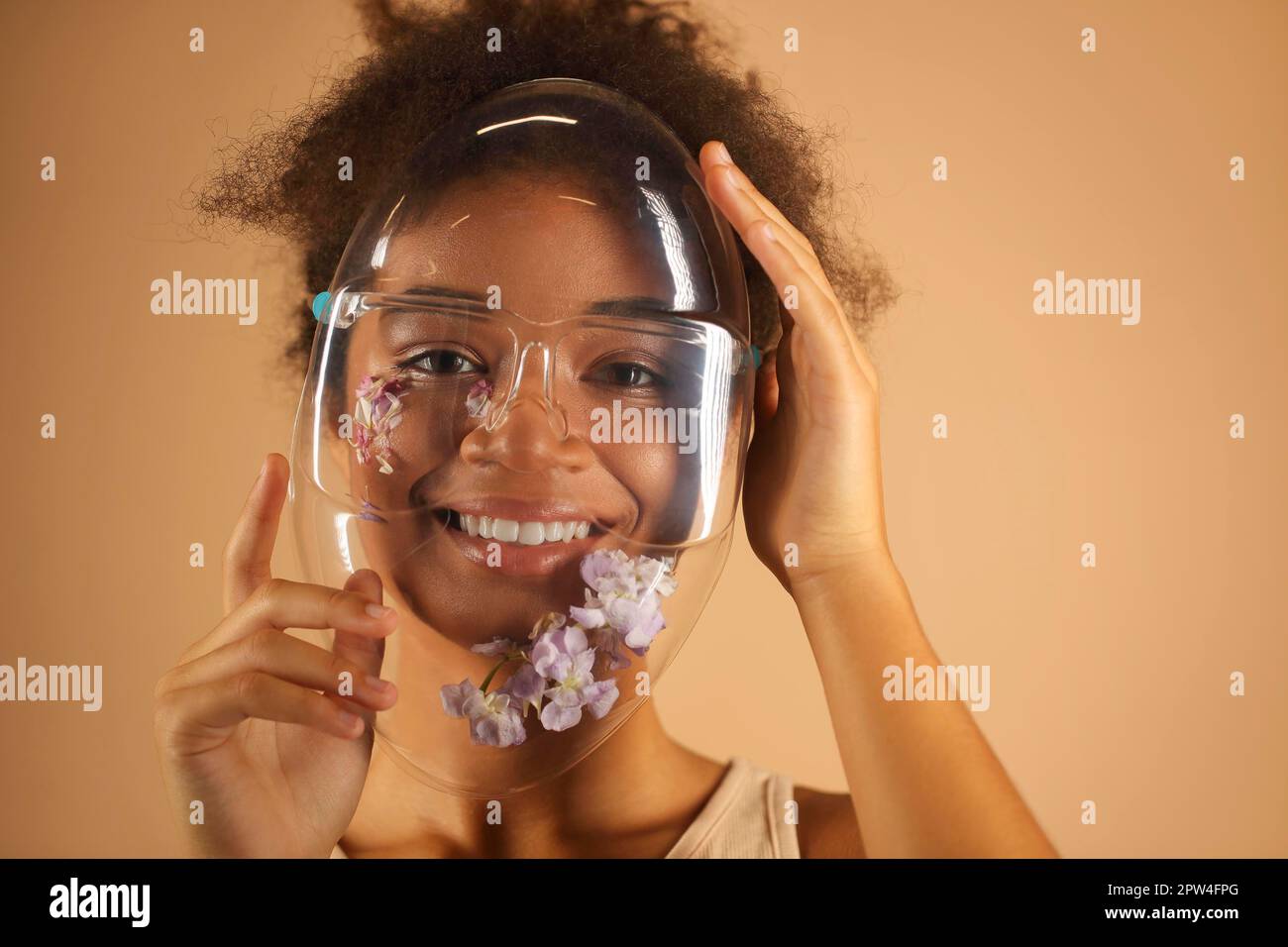 Jeune femme afro-américaine gaie portant un masque de protection en PVC transparent orné de fleurs et regardant la caméra avec des faisceaux Banque D'Images