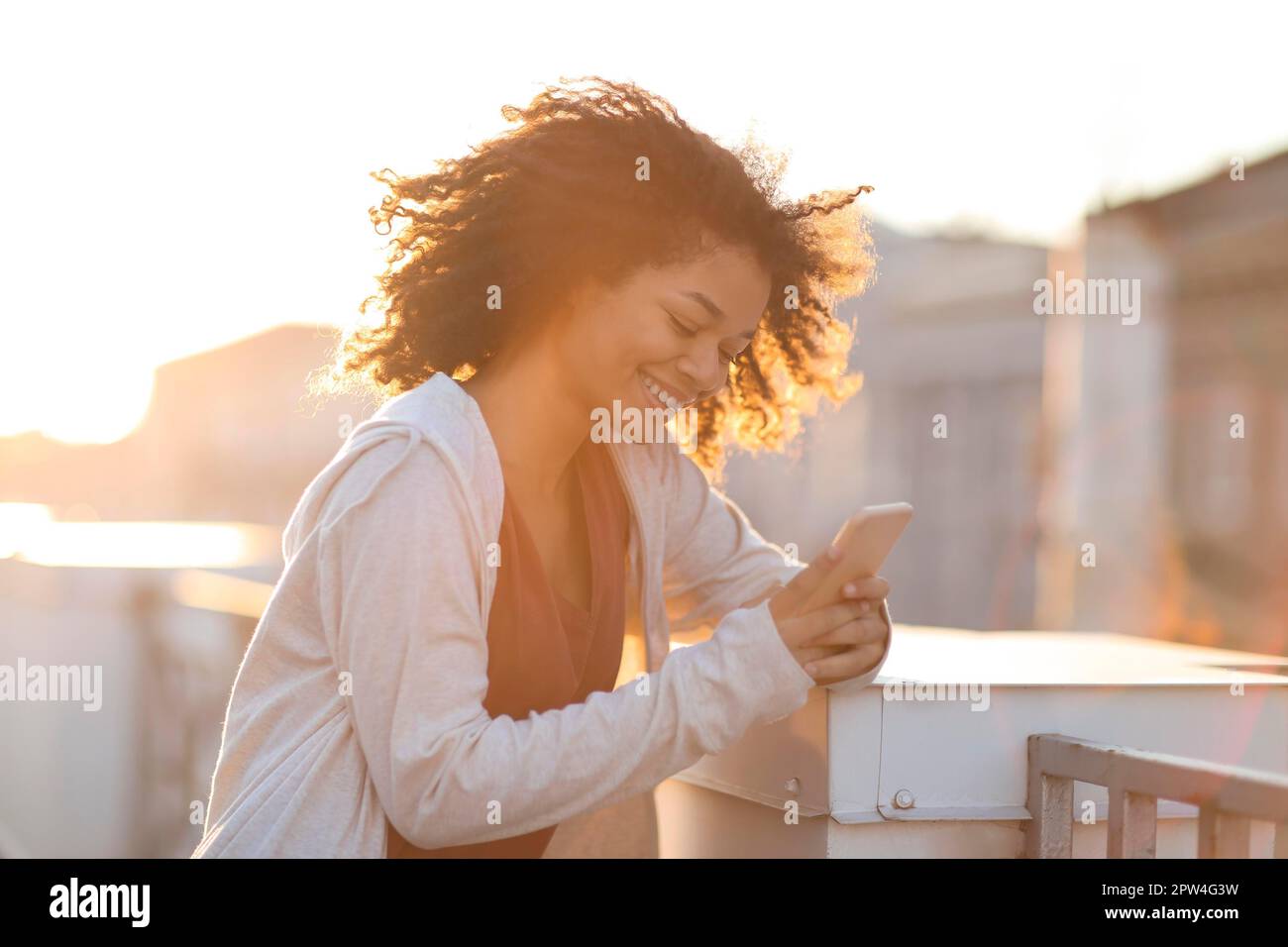 Portrait en plein air d'une femme afro-américaine joyeuse dans un casque à l'aide d'un smartphone et souriant heureux en se tenant sur le toit le matin Banque D'Images