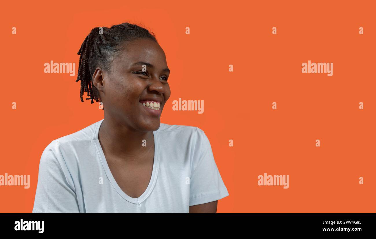 Portrait en gros plan de la jeune femme africaine heureuse regardant à droite avec un grand sourire, portant un t-shirt blanc et des cheveux de pain, sur fond orange avec Banque D'Images