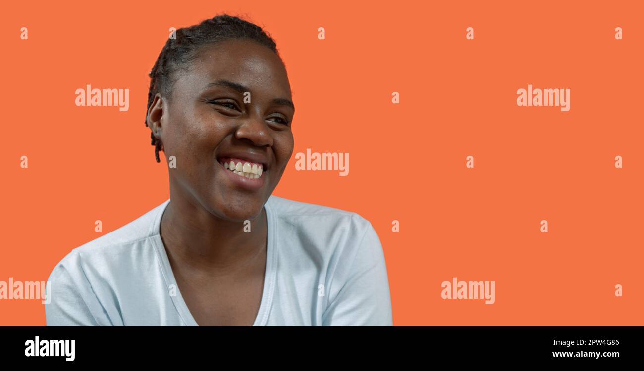 Portrait en gros plan de la jeune femme africaine heureuse regardant à droite avec un grand sourire, portant un t-shirt blanc et des cheveux de pain, sur fond orange avec Banque D'Images