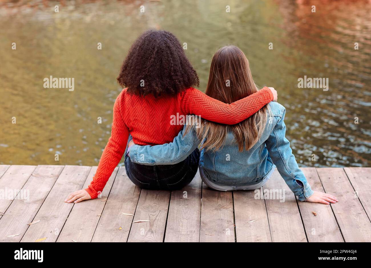 Vue arrière de deux amies multiraciales heureuses assises près de la rivière avec bonheur, deux meilleures amies de différentes races se détendant près de l'étang Banque D'Images