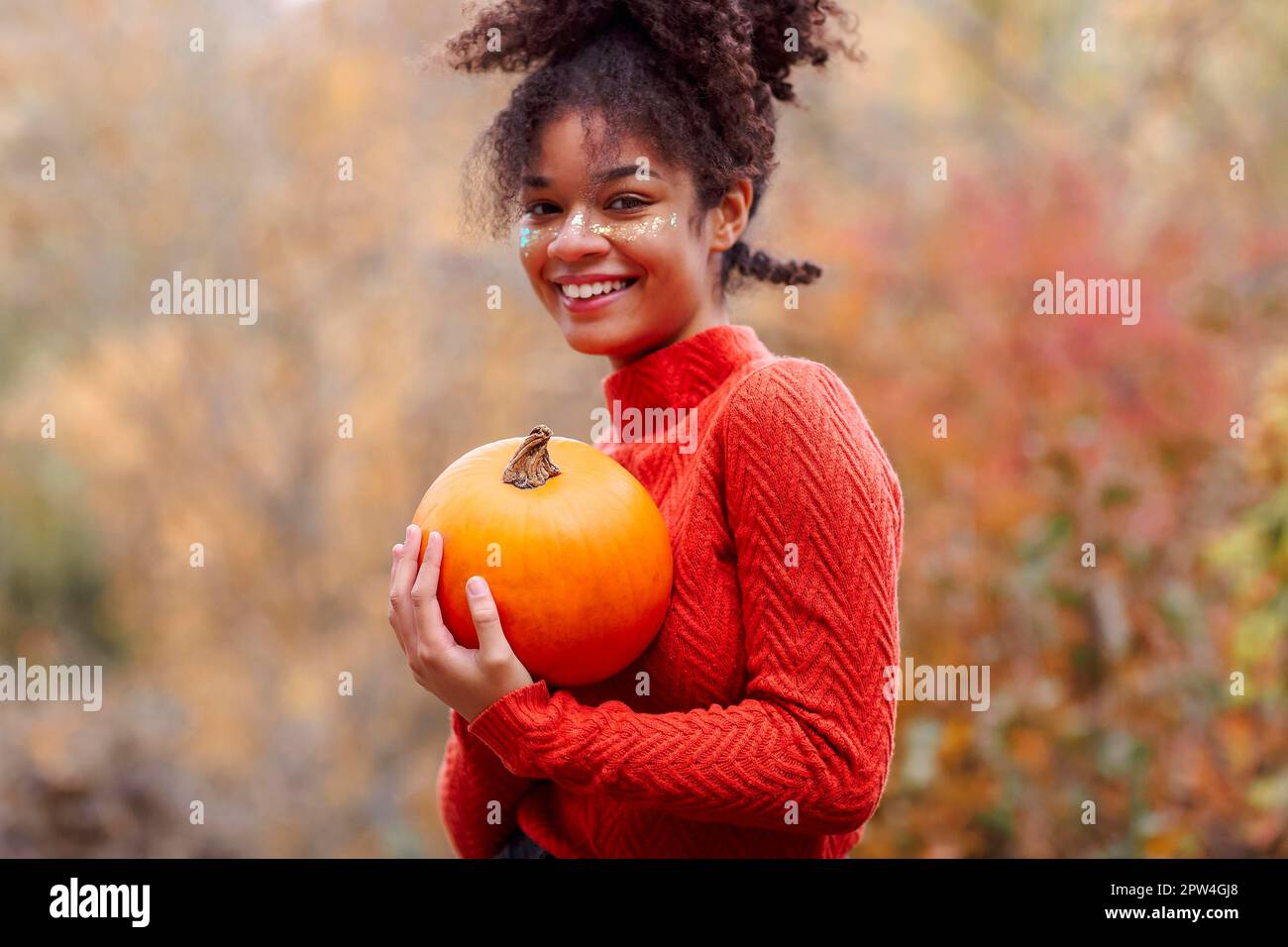 Jeune femme afro-américaine joyeuse avec des cheveux bouclés tenant la citrouille d'orange et souriant à la caméra tout en se tenant contre une forêt d'automne floue Banque D'Images