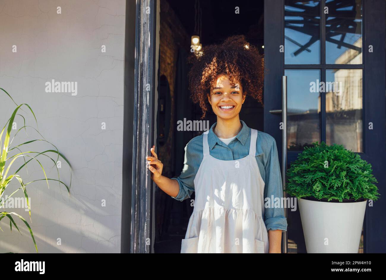 Propriétaire du café debout à l'entrée de son café. Joyeux jeune Barista jolie femme debout à la porte d'un restaurant et regardant la caméra Banque D'Images