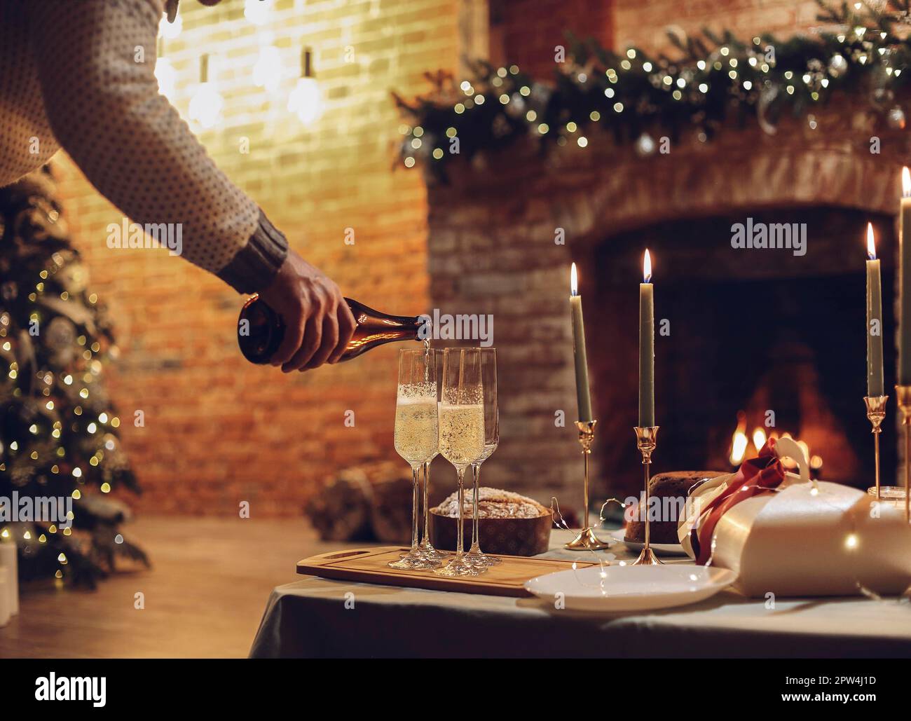 Célébration de la Saint-Sylvestre. Homme versant du champagne dans des verres debout sur la table avec un dîner de Noël festif, des bougies et des cadeaux enveloppés contre flou Banque D'Images