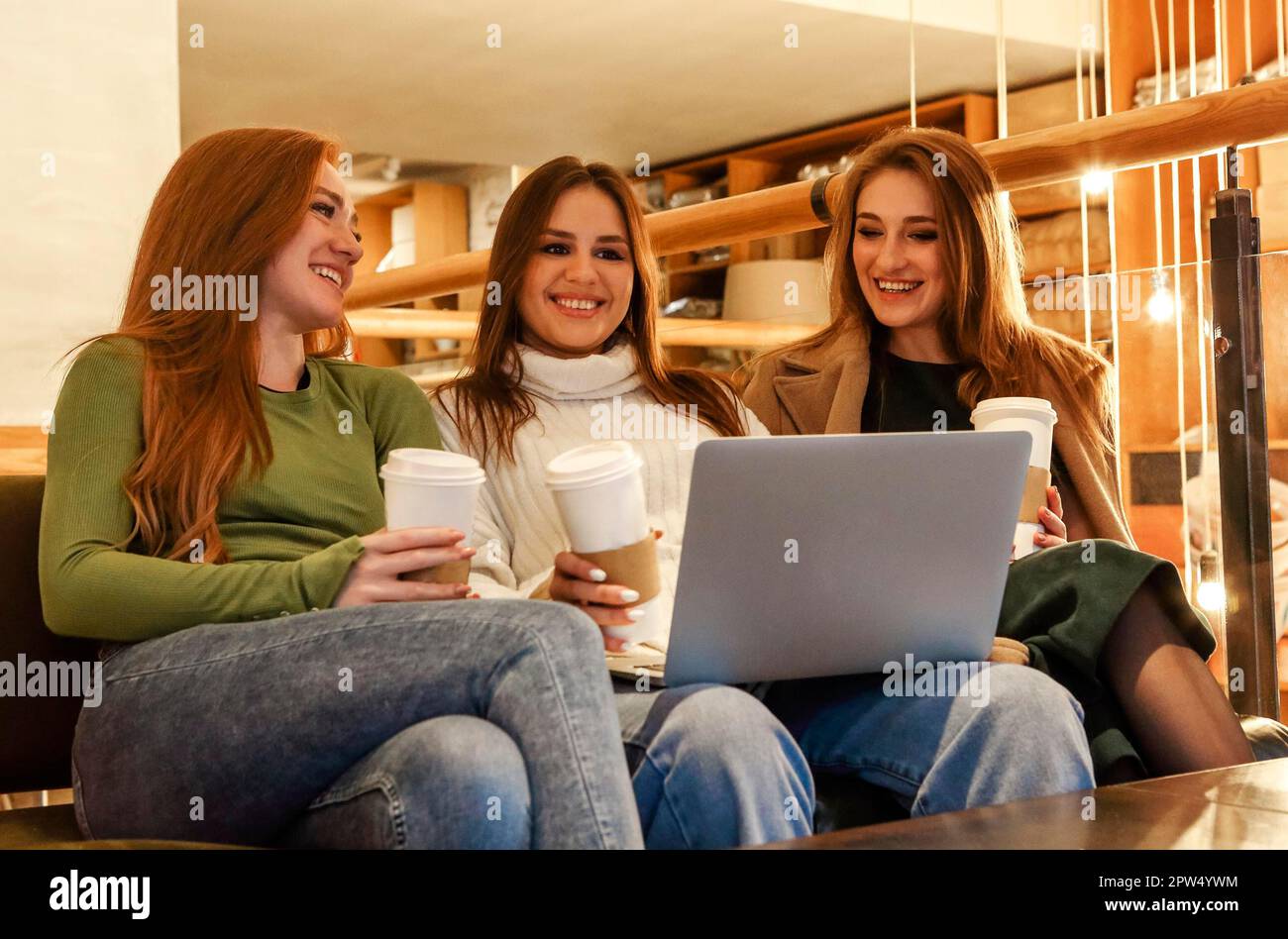 Des femmes gaies avec du café pour aller sourire et de naviguer sur ordinateur portable pendant que vous vous reposez à la cafétéria le week-end ensemble Banque D'Images