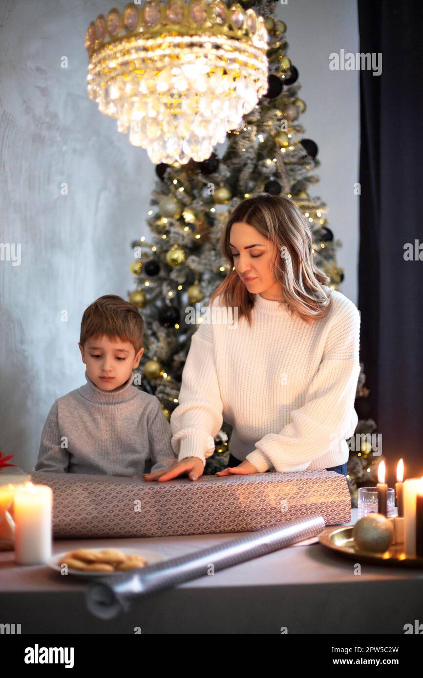 Mère et fils emballant les cadeaux de Noël ensemble à la maison tout en se tenant derrière la table près de l'arbre décoré, petit enfant aidant maman à emballer les vacances Banque D'Images