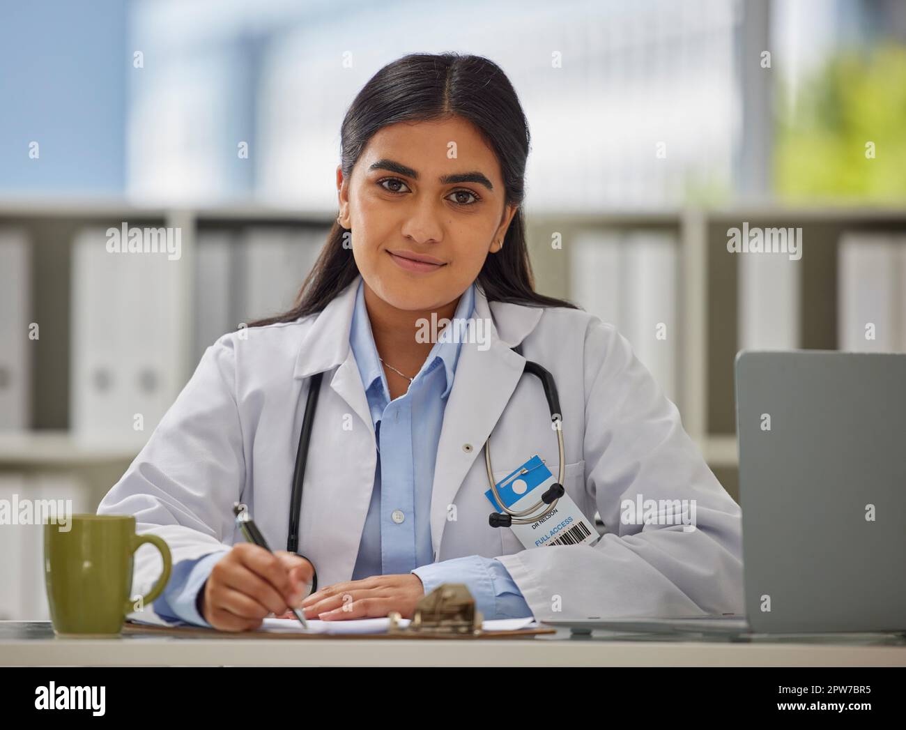 Portrait d'un jeune médecin indien portant un stéthoscope assis dans un bureau écrivant une ordonnance tout en étant assis à son bureau. Groupe de la cage mixte à l'aide d'un Banque D'Images