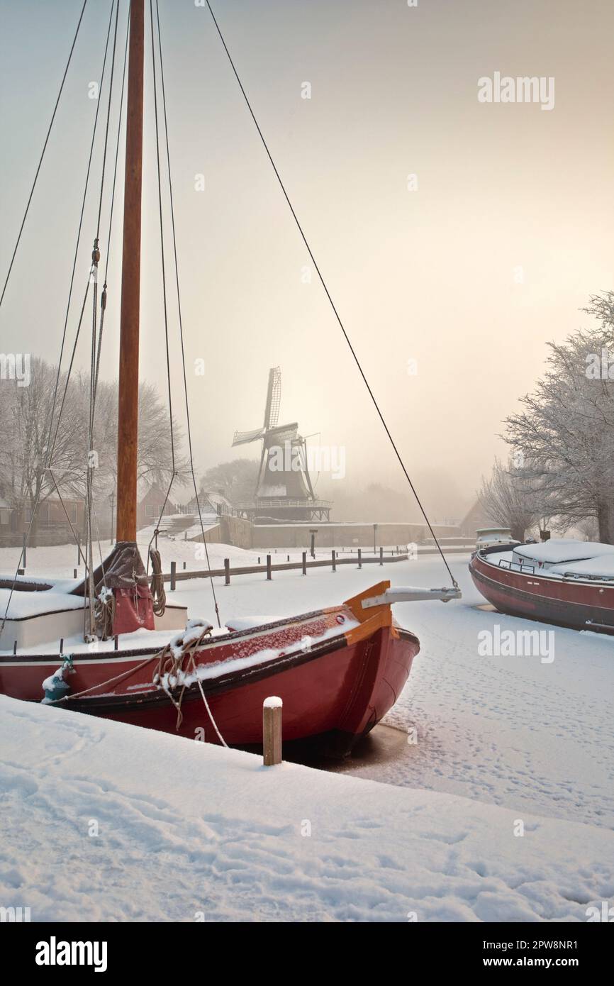 Pays-Bas, Sloten, navire traditionnel à voile dans le canal gelé. Moulin à vent de fond. Neige, hiver. Banque D'Images