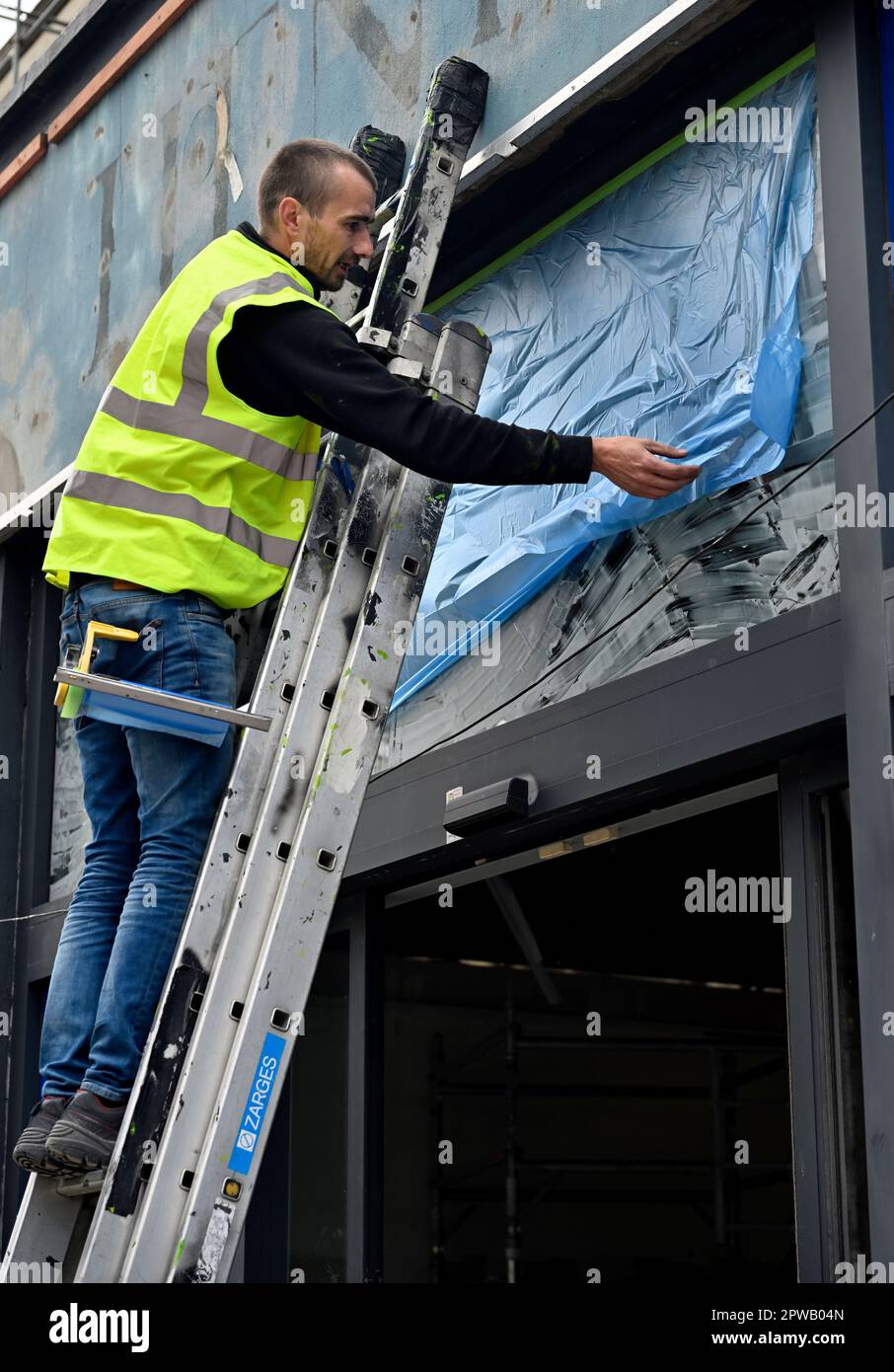 Workman sur l'échelle installer une feuille de masquage de protection en plastique sur la fenêtre en verre de l'atelier en vue de la peinture par pulvérisation Banque D'Images