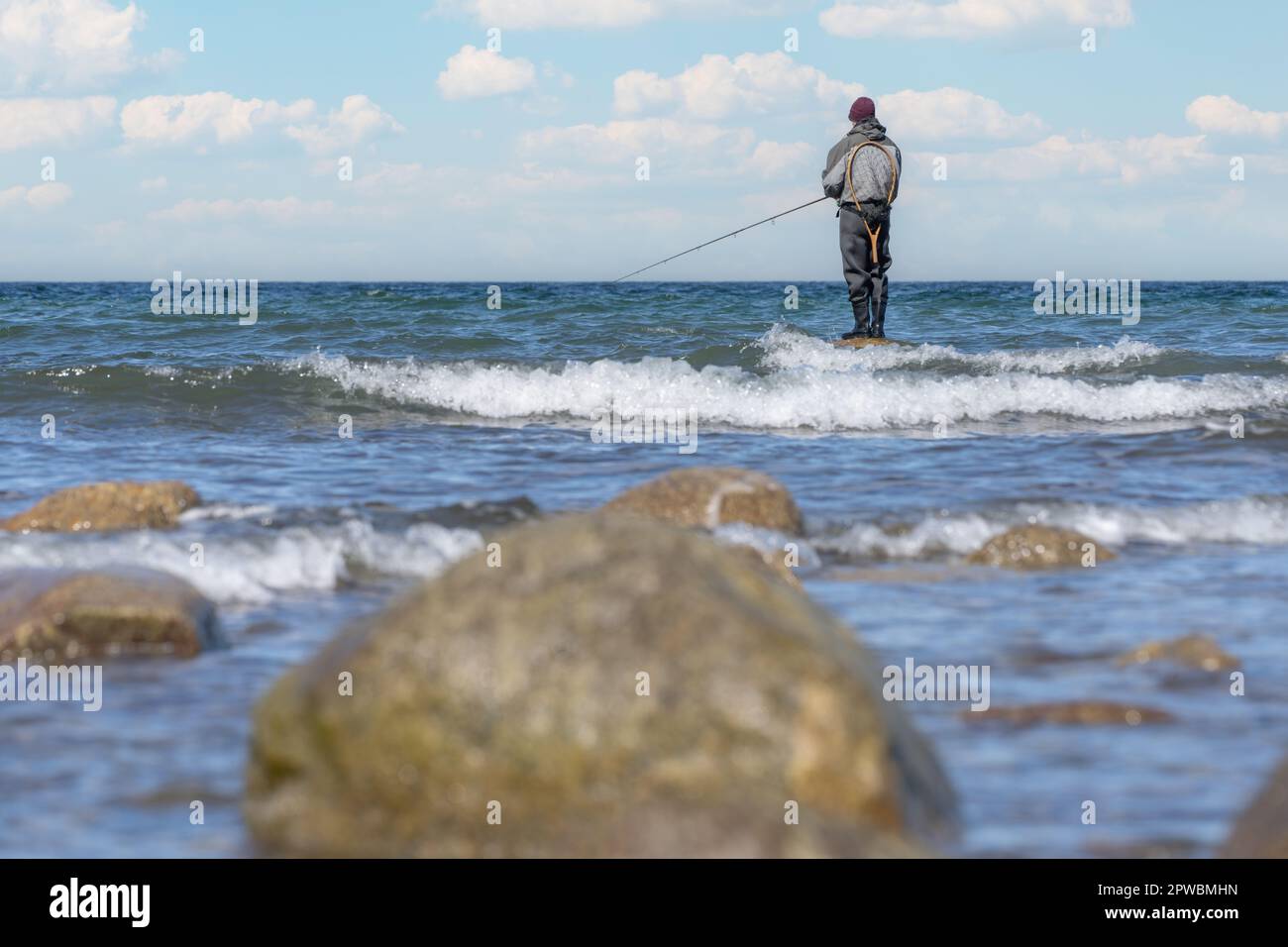 Un pêcheur à la ligne dans un pantalon imperméable pêche dans la mer Baltique. Il est debout sur une pierre dans l'eau - coucher de soleil sur la mer Baltique avec un pêcheur solitaire Banque D'Images