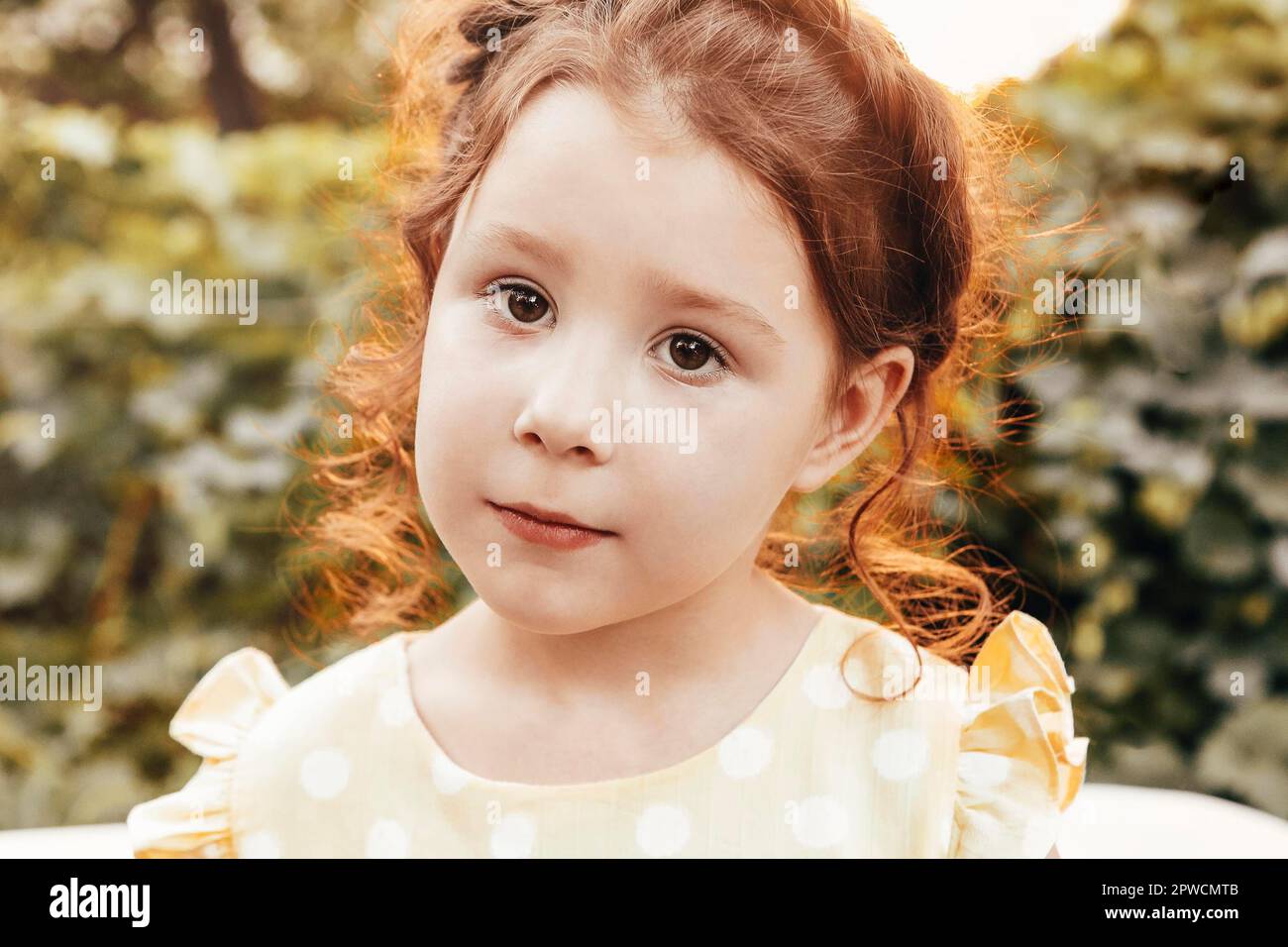 Portrait en gros plan de petite adorable fille à tête rouge avec cheveux bouclés et tresse portant une robe jaune à pois regardant l'appareil photo tout en jouant Banque D'Images