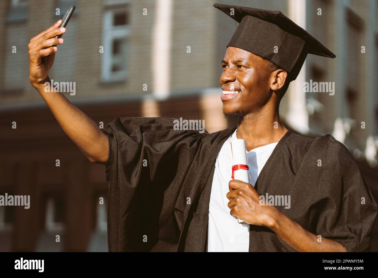 Un diplômé afro-américain souriant de l'université prend des photos avec l'appareil photo avant d'un smartphone avec un diplôme d'enseignement supérieur. Élève dans le manteau noir Banque D'Images