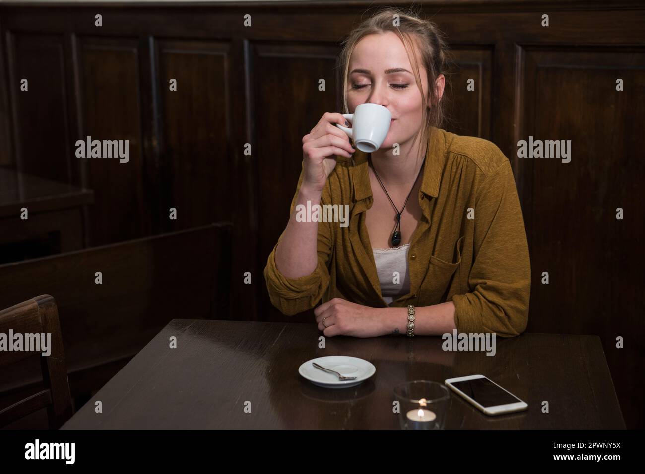 Jeune femme avec les yeux fermés buvant du café tout en mobile gardé sur la table au restaurant Banque D'Images