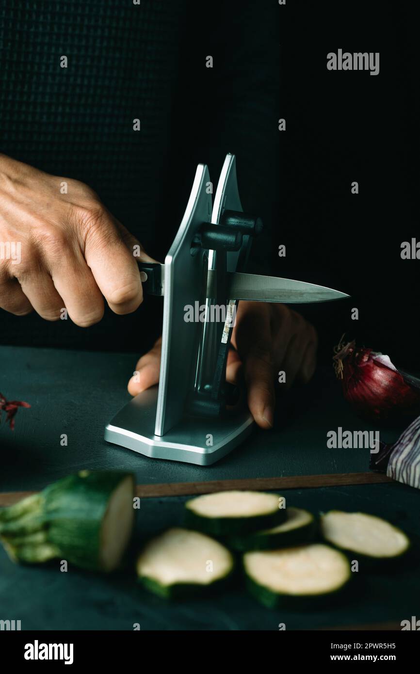 gros plan d'un homme aiguisant un couteau de cuisine avec un taille-crayon de bureau placé sur un à côté de quelques légumes hachés Banque D'Images