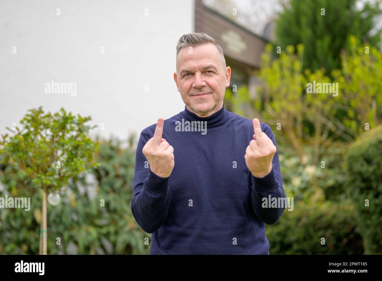Homme d'âge moyen avec cheveux gris, non rasé, avec un chandail bleu à col montant se tient dans le jardin et montre les deux doigts du milieu dans l'appareil photo Banque D'Images