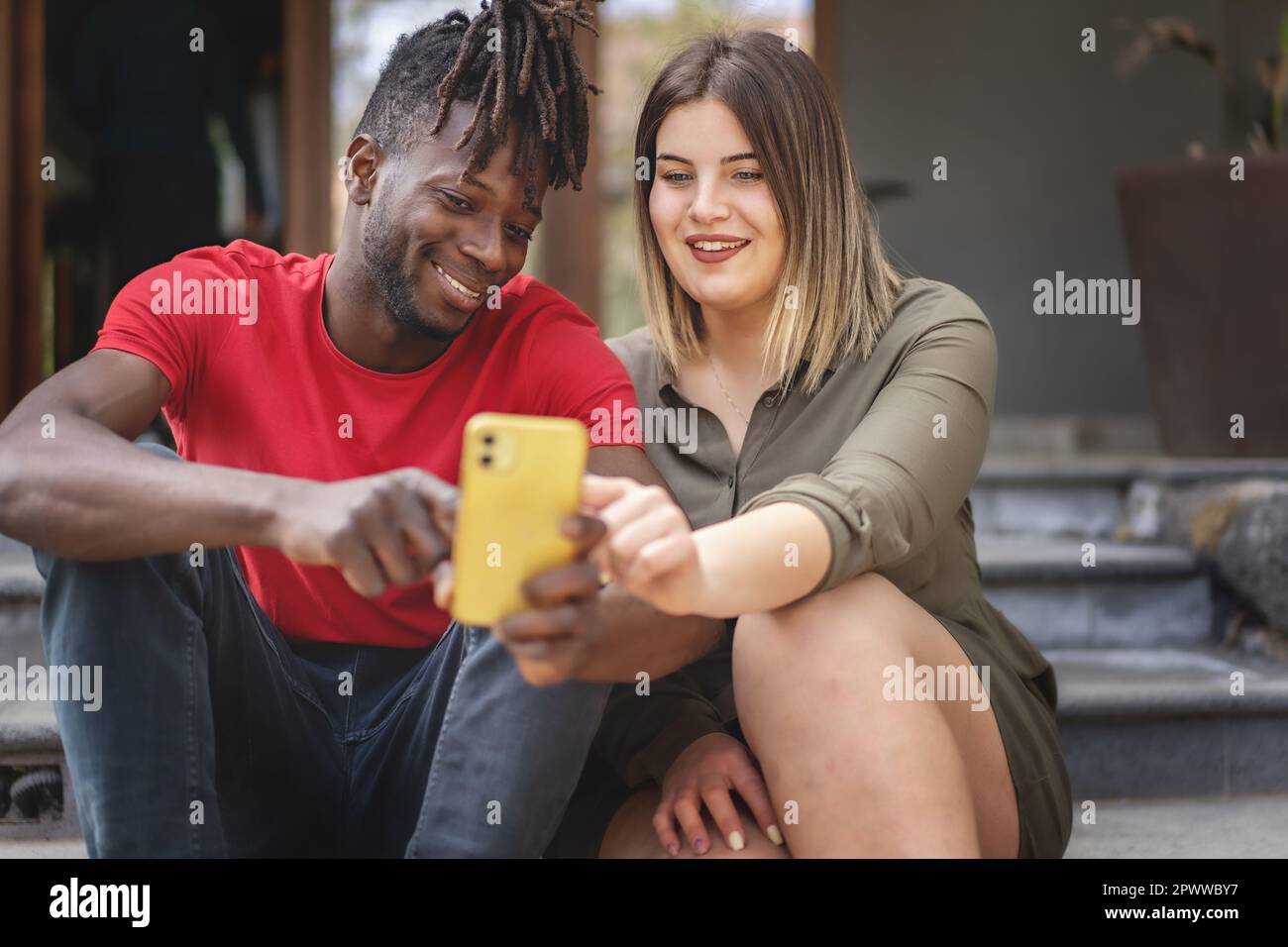 Deux amis divers, un Africain et une femme de taille plus, assis sur des escaliers extérieurs, regardant un contenu de smartphone lors d'une journée ensoleillée d'été. Banque D'Images