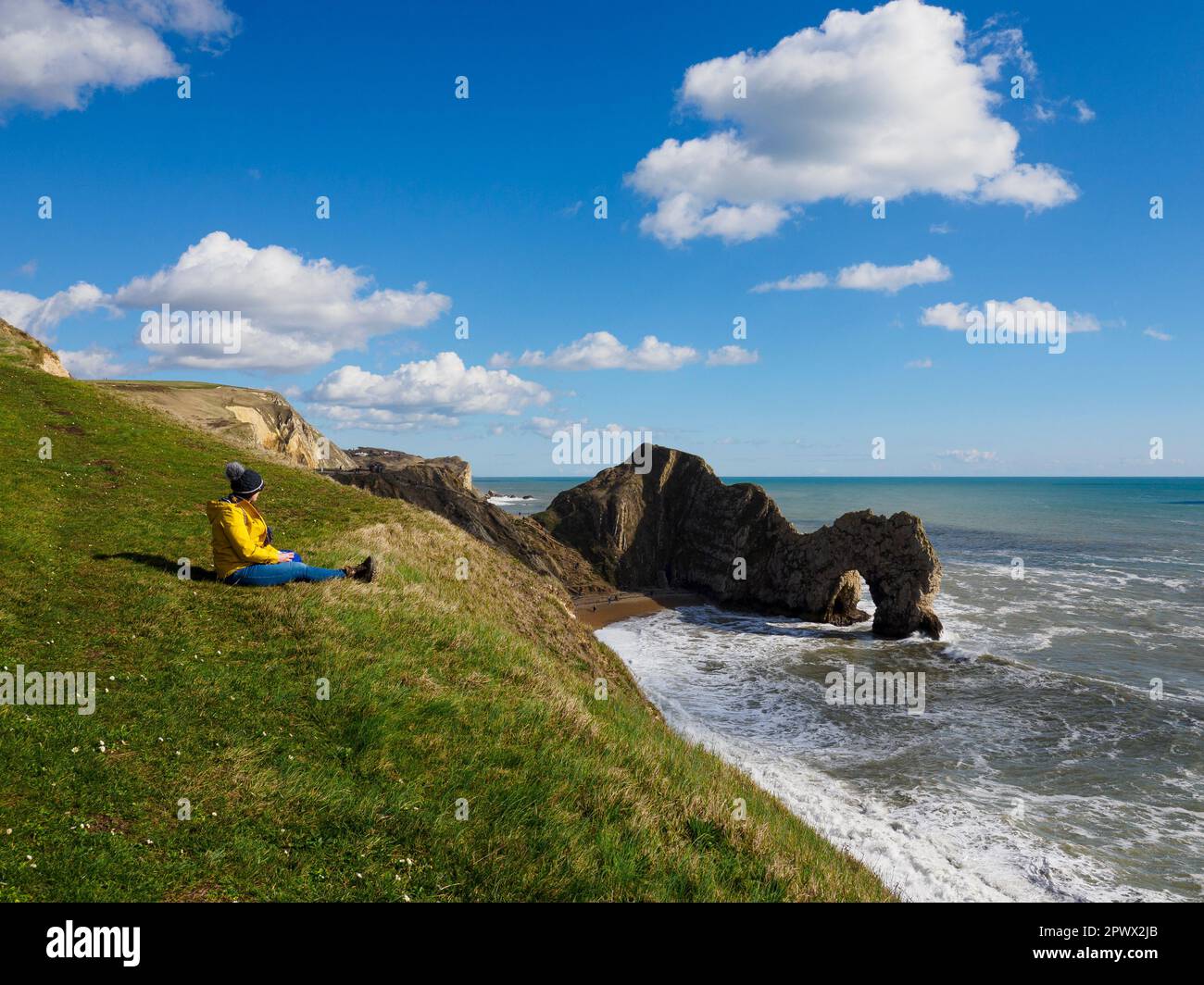 Femme assise sur une falaise au-dessus de la porte et de la plage de Durdle lors d'une journée ensoleillée en mars, Dorset, Royaume-Uni Banque D'Images