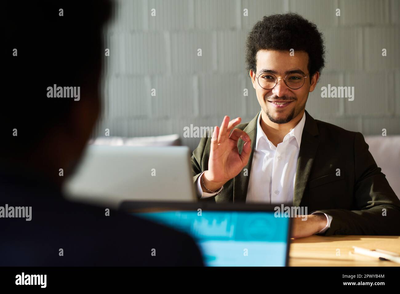 Jeune homme d'affaires qui se remplit montrant un signe correct à son collègue ou candidat avec un ordinateur portable assis devant lui pendant la communication Banque D'Images