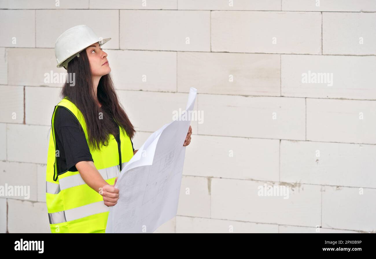 Jeune femme en casque blanc à chapeau rigide et gilet jaune vert haute visibilité, debout à côté du mur de construction, tenant des plans de papier de plans, lookin Banque D'Images
