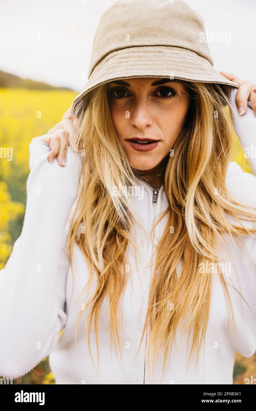 Belle jeune femme blonde, portant un chapeau, pose au milieu d'un champ de fleurs de colza jaune Banque D'Images
