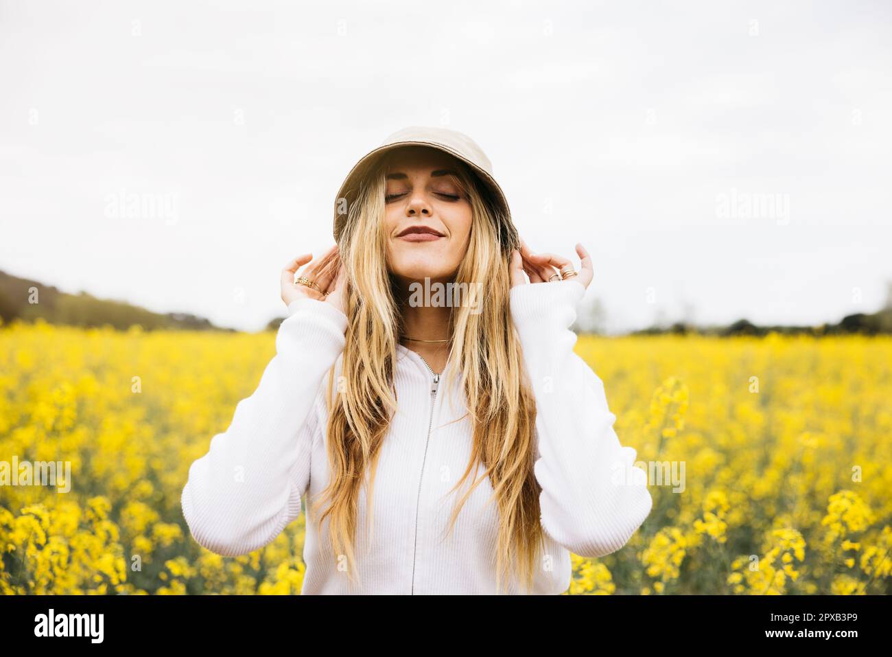 Belle jeune blonde gaie femme, portant un chapeau, pose au milieu d'un champ de fleurs de colza jaune Banque D'Images