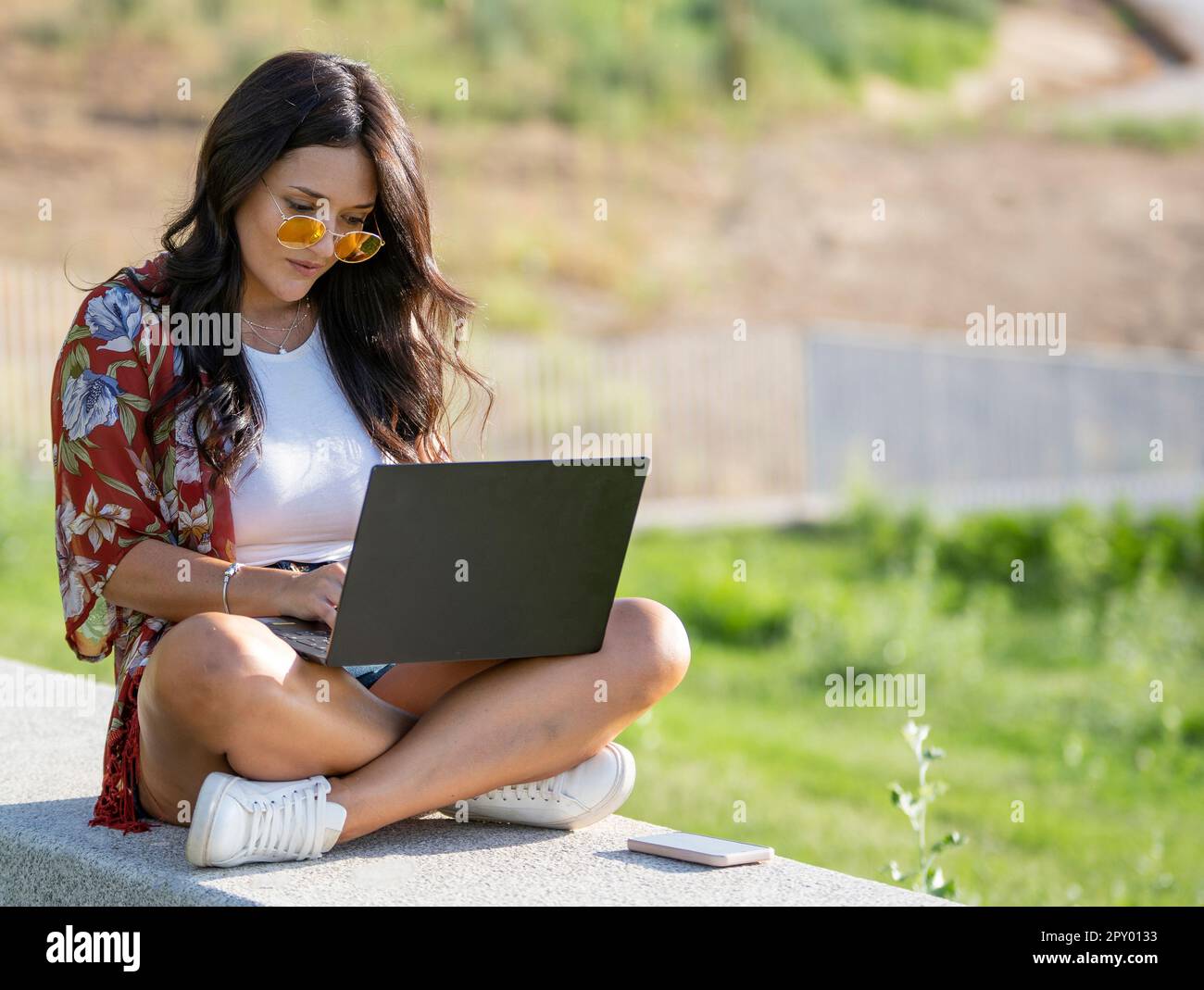 Jeune femme en lunettes de soleil à l'aide d'un ordinateur portable tout en étant assise sur un banc dans un parc. Concept de technologie. Banque D'Images