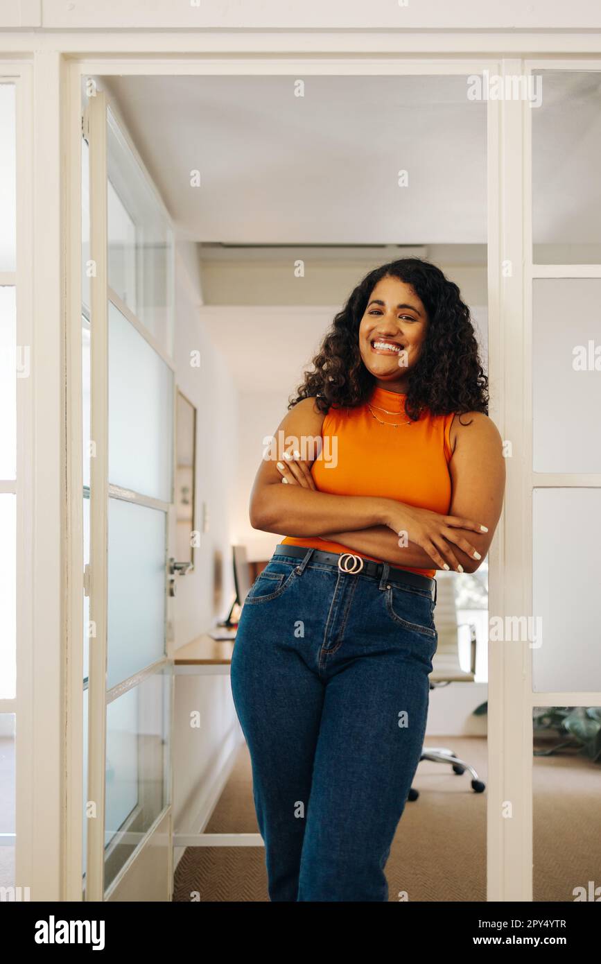 Une femme entrepreneure heureuse souriant à la caméra tout en se tenant dans un bureau avec ses bras croisés. Femme d'affaires réussie travaillant dans un travail créatif Banque D'Images
