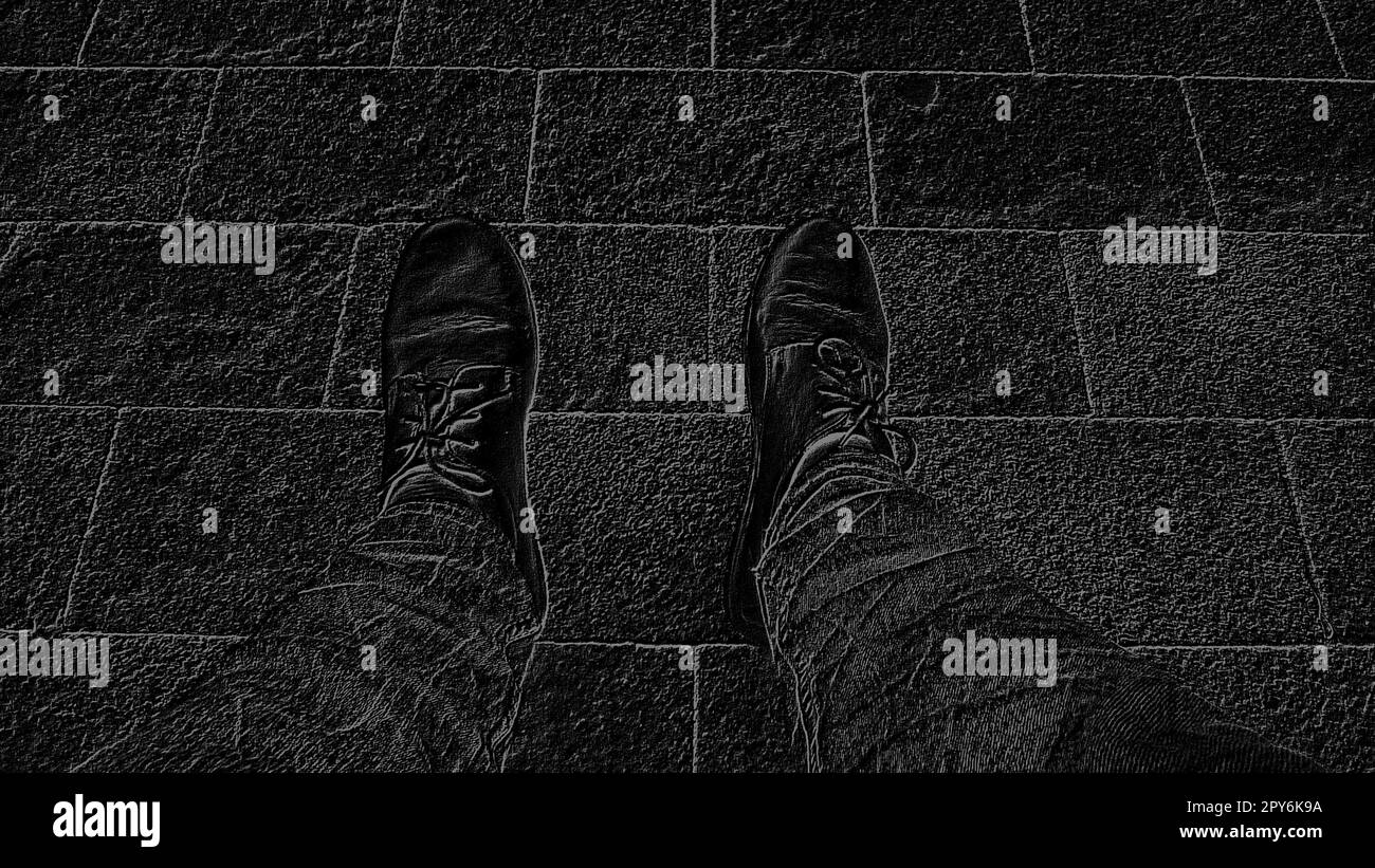 Image monochrome en noir et blanc. Pieds dans les bottes contre le fond de tuiles de rue ou de la route. Deux pattes d'un homme assis, photographiées d'en haut tout en étant assis. Banque D'Images