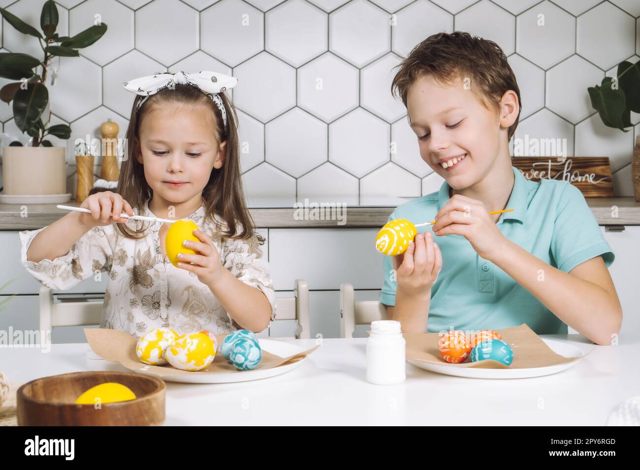 Portrait de sourire joyeux deux enfants petite fille, garçon, peinture, coloration des oeufs de Pâques, table de cuisine avec des pinceaux. Banque D'Images