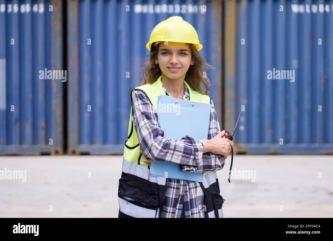 Jeune femme caucasienne avec gilet de sécurité et support de casque jaune avec le bras plié tout en tenant le presse-papiers et le talkie-walkie. Un grand conteneur de fret est en arrière-plan Banque D'Images