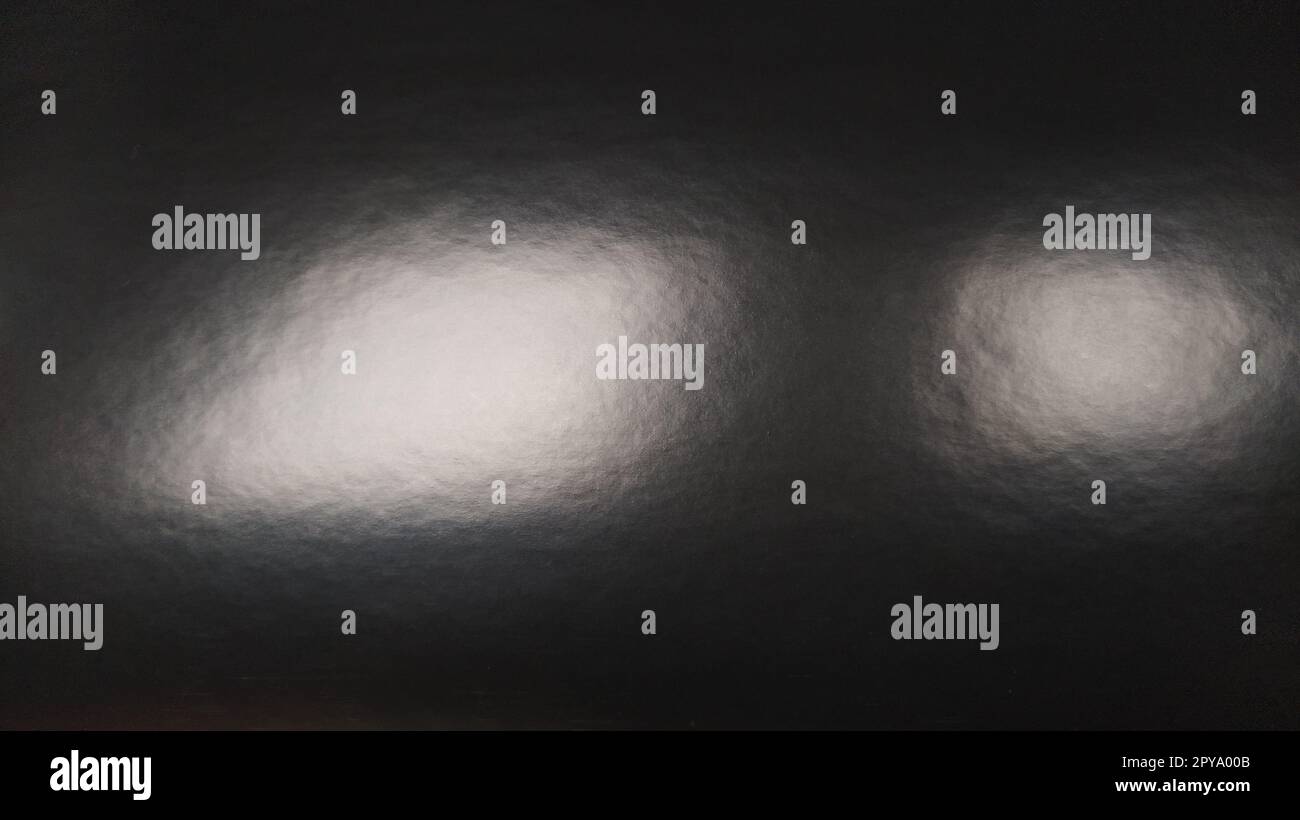 Éblouissement de lumière sur papier gris brillant. Plusieurs taches lumineuses sur une surface sombre qui réfléchit la lumière. Fond monochrome abstrait. Banque D'Images