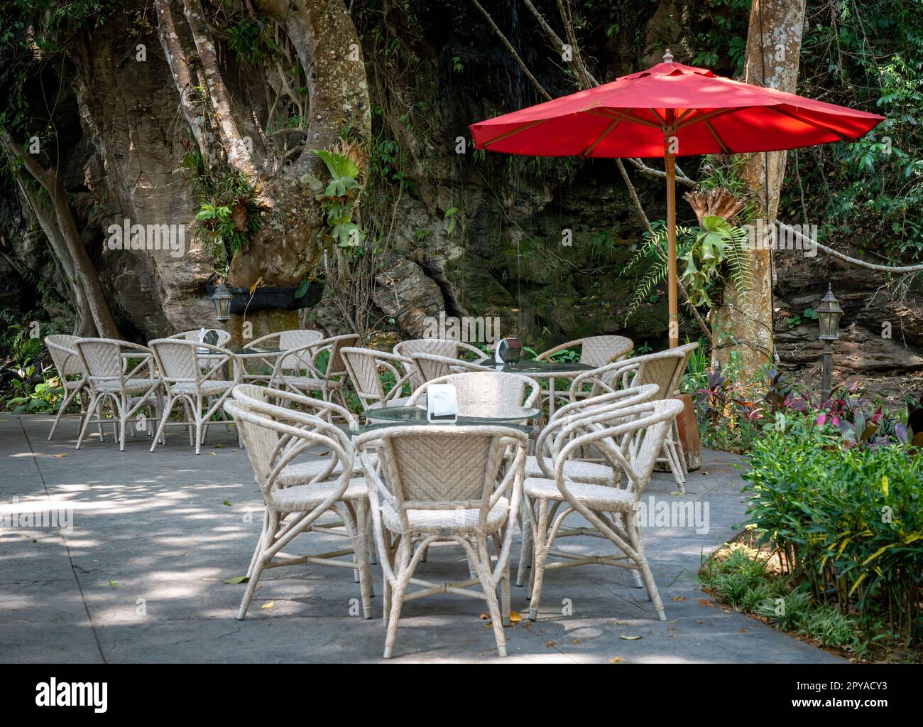 Un café en plein air, avec plusieurs tables et chaises disposées autour d'un parapluie rouge vif, sur fond d'arbres verts luxuriants Banque D'Images